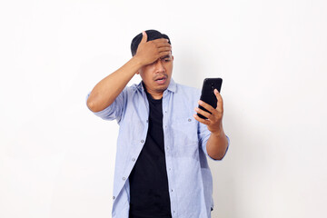 Young Asian man using smartphone stressed with hand on head, shocked with shame and surprise face, angry and frustrated over white wall background. Fear and upset for mistake.