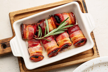 Baking dish with delicious prunes baked in crispy bacon and rosemary branch on light background