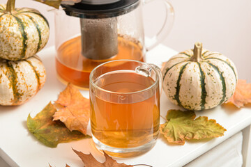 Tea pot, glass cup of hot beverage, pumpkins and autumn leaves on table near beige wall