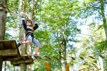 Naklejka na ściany i meble School boy in forest adventure park. Acitve child, kid in helmet climbs on high rope trail. Agility skills and climbing outdoor amusement center for children. Outdoors activity for kid and families.