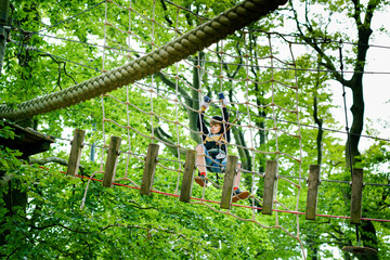 Naklejka na ściany i meble School boy in forest adventure park. Acitve child, kid in helmet climbs on high rope trail. Agility skills and climbing outdoor amusement center for children. Outdoors activity for kid and families.