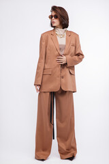 Fashion photo of a beautiful elegant young woman in a pretty oversize brown and beige suit, corset, stylish sunglasses, jacket, pants posing over white background. Bob haircut. Studio Shot. Portrait
