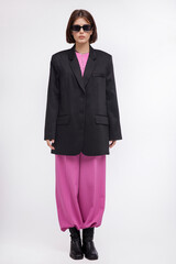 Fototapeta premium Fashion photo of a beautiful elegant young woman in a pretty oversize black jacket and pink suit, boots, stylish sunglasses posing over white background. Bob haircut. Studio Shot