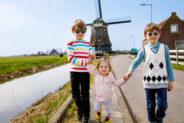 Two school kids boys and little baby girl walking on street with big windmill in Netherlands....