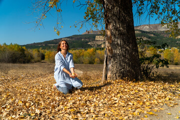 Smiling woman in blue long shirt sitting under a tree and tossing up autumn leaves