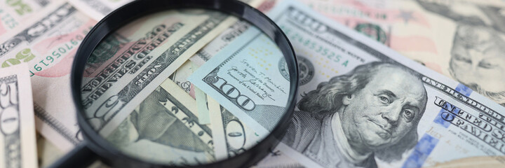One hundred dollar bills and magnifying glass lie on table closeup