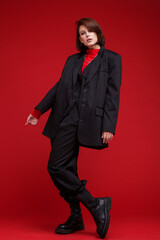 Fashion photo of a beautiful elegant young woman in a pretty oversize black suit, pants, top posing over red background. Bob haircut. Studio Shot. Rebellious free street style. Rebel and classic