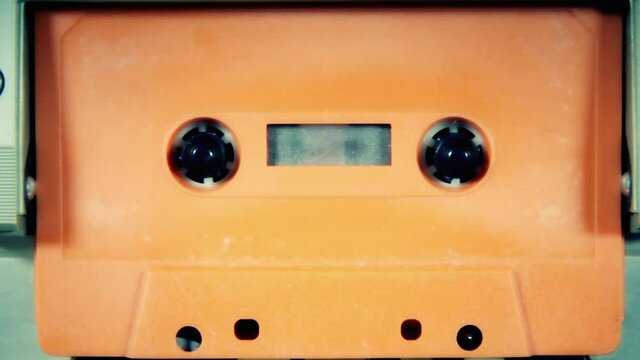 Inserting an orange cassette tape (not labeled) into a deck and playing its content. Retro vintage old magnetic media, nostalgic vibes. Front shot.
