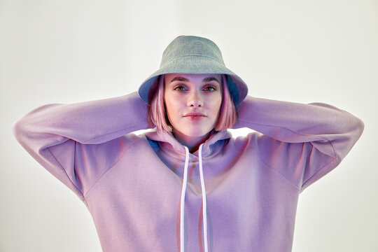 Self assured young female millennial with pink hair in trendy panama hat and hoodie touching head and looking at camera against white background