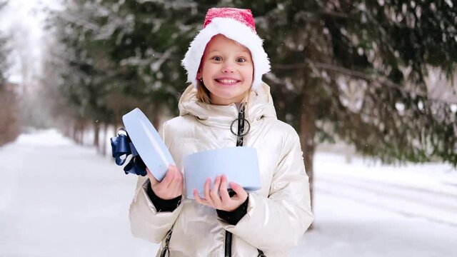 Little girl in red christmas hat opening blue gift box and being amazed, surprised, shocked. Happy child in Santa hat enjoying present in winter forest. Outdoors winter activities.