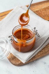 Homemade salted caramel sauce in glass jar on wooden board, grey background. Caramel sauce drips...