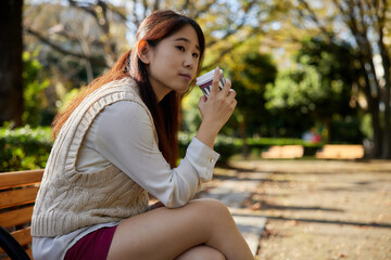 Fototapeta premium コーヒーカップを持ってベンチに座っているアジア人女性