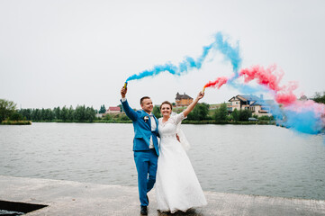 Bride and groom holding blue and red colored smoke bombs on nature. Newlyweds with smoke bombs...