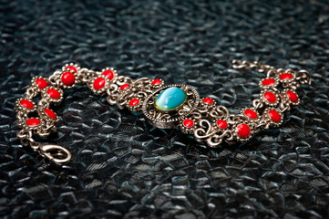 Metal women's wrist bracelet with an emerald and red stones on a dark background.
