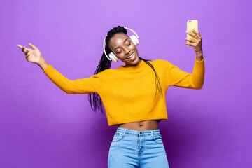 African American woman wearing headphones listening to music from smart phone and dancing on colorful purple isolated studio background