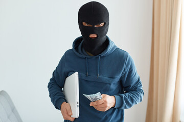 Indoor shot of burglar robbed the house and leaves with the stolen laptop and money, male burglar...