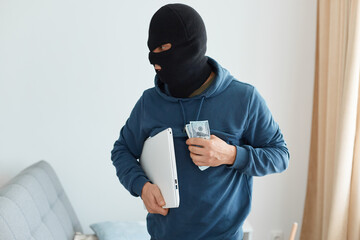 Indoor shot of male burglar wearing dark blue hoodie and black mask holding portable computer and...