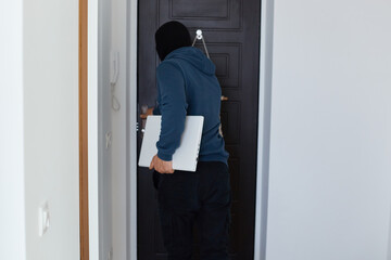 Back view of man robber wearing blue hoodie and black balaclava standing with notebook he stole in hands, leaving place of crime, stole other people's personal belongings.