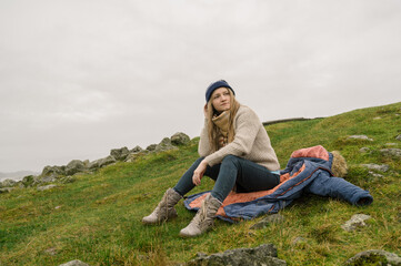 Young beautiful woman in knitted sweater and hat is sitting on her coat on ground. Cloudy day. Scandinavian nature.