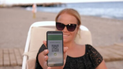 Portrait of a young woman on the beach in a swimsuit and sunglasses, she is holding a phone with a QR code about vaccination against coronavirus infection. - Powered by Adobe
