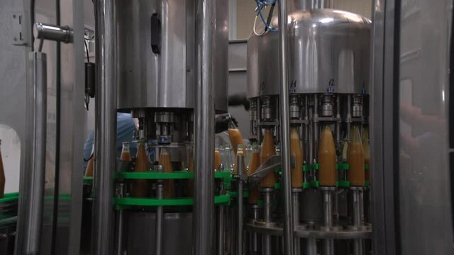 Close up inside of mechanism bottling line. Orange-colored sea-buckthorn juice in glass bottles. Worker on background change some bottles