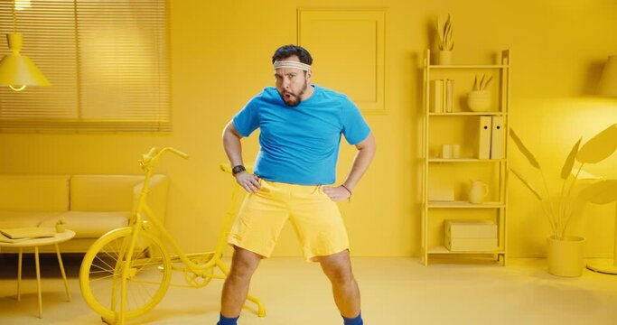 Slow motion of funny handsome young man in blue t-shirt doing aerobics with silly movement at home with yellow interior. Bearded male model in sportswear having a workout with funny expression