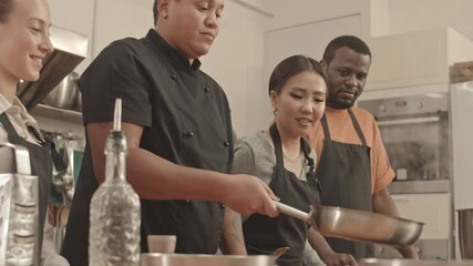 Low angle of young female chef flipping pieces of chicken fillet in pan, Asian and Caucasian women and African American man standing on her sides in large kitchen, watching