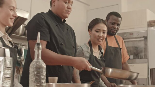 Low angle of young female chef flipping pieces of chicken fillet in pan, Asian and Caucasian women and African American man standing on her sides in large kitchen, watching