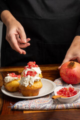 Top view of woman's hands holding a pomegranate, three muffins with cream cheese and pomegranate seeds on wooden table, selective focus, vertical