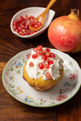 Top view of homemade muffin with cream cheese and pomegranate seeds, selective focus, on white plate, on wooden table, with bowl of seeds and pomegranate, vertical
