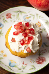 Aerial view of homemade muffin with cream cheese and pomegranate seeds, selective focus, in white flower plate, on wooden table, vertical