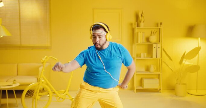 Excited bearded man in casual clothes listening music on mobile phone apps while wearing headphones and dancing at home in a room with yellow interior. Joyful young man enjoying a song on smart phone