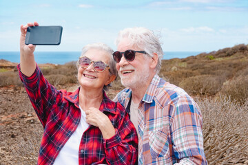 Attractive carefree senior couple in outdoors excursion enjoying selfie with mobile phone wearing checkered shirts.  Horizon over sea
