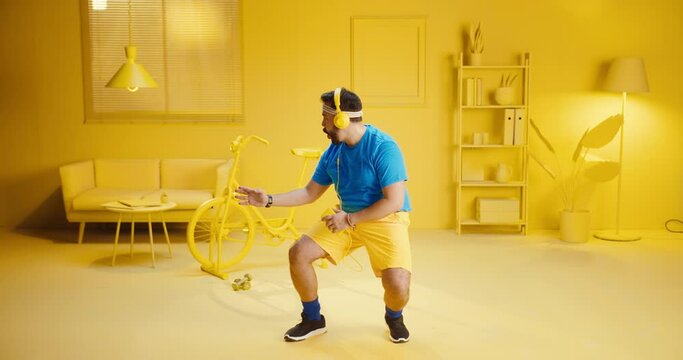 Handsome bearded young man playing music on mobile phone app and start dancing or exercise in a room with monochrome yellow color interior. Man listening workout music and dancing at home