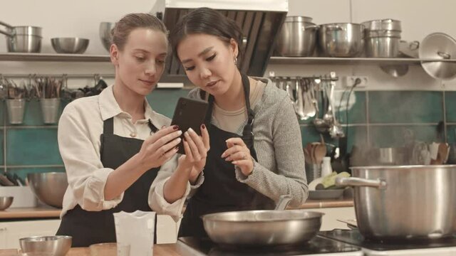 Medium low angle of young Asian and Caucasian women wearing aprons, standing by stove in professional kitchen, using smartphone for recipe, talking and smiling