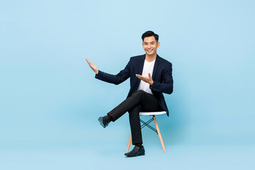 Smiling young Asian man sitting cross legged with hands open to empty space aside in light blue isolated background - obrazy, fototapety, plakaty