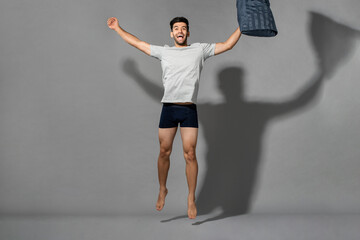 Fresh and energetic young Caucasian man in sleepwear jumping with pillow after wake up in light gray isolated background, good sleep hygiene concept