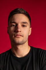 Fototapeta premium Close-up portrait of young handsome man in black t-shirt looking at camera isolated on dark red studio background.