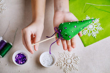 Decorative Christmas tree made of green felt with purple sequins and white beads. Crafts with children at school and at home. The child is holding a Christmas toy. - Powered by Adobe