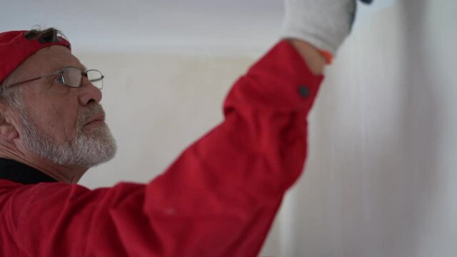 Gray-bearded man with a tassel in a red cap makes repairs in his new home. Retiree paints the wall with white paint