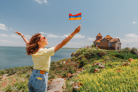 Happy tourist woman with armenian flag posing at the Hayravank Monastery on the shore of Lake Sevan.