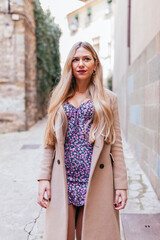Confident blond latin stylish woman wearing beige coat and floral dress walking on the street durinng winter. Cheerful argentinian female walking in the city.