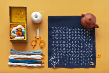 Handmade geometric embroidery in style of Japanese Sashiko with white threads on blue cotton fabric and accessories for embroidery on orange background. Teapot with green tea nearby. Flat lay, closeup