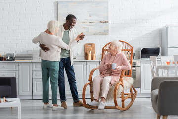Smiling senior woman holding cup in rocking chair near interracial friends dancing in nursing home