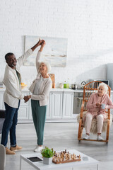 Smiling multiethnic pensioners dancing near friend with tea in nursing home