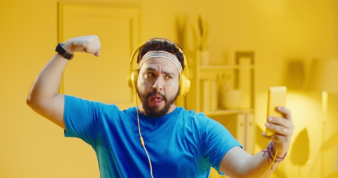 Slow motion portrait of carefree young bearded man wearing headphones and taking selfie picture with a smart phone while showing his arms muscle at home with monochrome yellow interior.