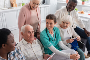 Fototapeta na wymiar Senior man holding digital tablet near nurse and interracial friends in nursing home