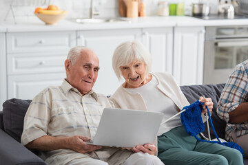 Obraz premium Senior man holding laptop near cheerful friend with yarn in nursing home