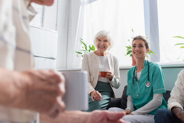 Positive woman with tea and nurse looking at senior man in nursing home