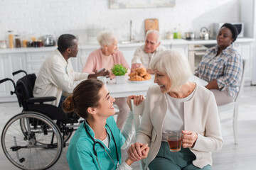 Fototapeta premium Positive nurse looking at patient with tea near blurred elderly people in nursing home